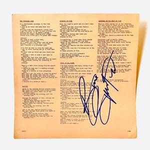 Puede incluir: Una hoja cuadrada de papel color canela claro con letras de canciones impresas en negro, incluyendo The Promised Land, Streets of Fire y Darkness on the Edge of Town. Una firma grande y audaz en tinta azul aparece en la parte inferior derecha.