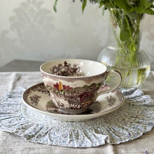 Puede incluir: Un juego de taza y platillo vintage con un diseño de paisaje marrón y blanco. La taza y el platillo se colocan sobre un mantel individual con estampado floral. Un jarrón de vidrio transparente con tallos verdes y flores moradas está en el fondo.