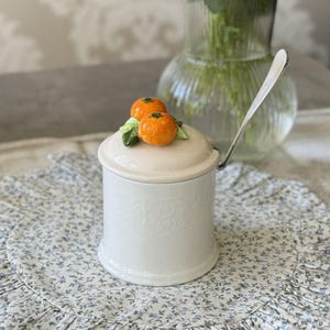 Puede incluir: Un tarro de cerámica blanco con tapa, adornado con dos frutas naranjas y hojas verdes. El tarro tiene una cuchara plateada y está colocado sobre un mantel individual con estampado floral. El tarro tiene un diseño en relieve.