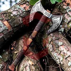 May include: A double-bit axe featuring a dark metal head with engraved white wing motifs and a central Celtic knot design. The handle is constructed from wood with a dark brown leather wrap, featuring a ribbed texture and a flared base at the end.