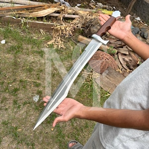 May include: A silver and grey sword with a wooden handle. The blade is long and curved, with a sharp point. The handle is brown and has a carved design. The sword is held in front of a grassy background.
