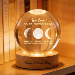 May include: A lit crystal sphere on a wooden base, with etched moon phases and the text "We Love YOU TO THE MOON & BACK." The base is engraved with "Happy Mother's Day." The sphere is surrounded by books on a wooden shelf.