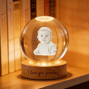 May include: A clear crystal ball with a baby's image inside, resting on a wooden base. The base has the inscription "I love you forever" and is illuminated by a warm light. The item is displayed on a wooden shelf with books.