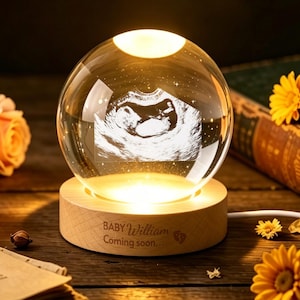 May include: A lit crystal ball lamp displaying an ultrasound image, set on a wooden base. The base is engraved with the text "BABY William Coming soon." The scene includes orange roses and yellow flowers, arranged on a wooden surface.