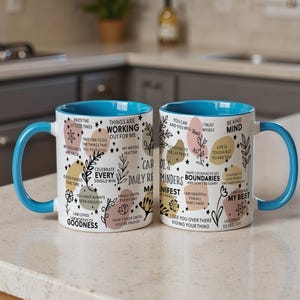 May include: Two white ceramic mugs with blue handles and rims. The mugs are decorated with inspirational phrases, floral designs, and colorful shapes. The text includes phrases like "Things are working out for me" and "Celebrate every single win."