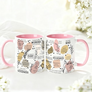 May include: Two white ceramic mugs with pink handles and interiors. The mugs are decorated with inspirational phrases and floral designs in black, gold, and pink. The text includes phrases like "Be Kind Mind" and "Celebrate Every Single Win."