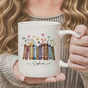 以下が含まれることがあります： 白いセラミックマグカップ。花と蝶のデザインが本の上に描かれています。マグカップには筆記体で「Sophia」と書かれています。本には「CUSTOM TEXT」と書かれています。
