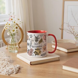 May include: A white ceramic mug with a red interior and handle, featuring the text "Becky's Daily Reminders" and other affirmations. The mug is decorated with floral and abstract designs. It sits on a cream-colored notebook on a wooden table.