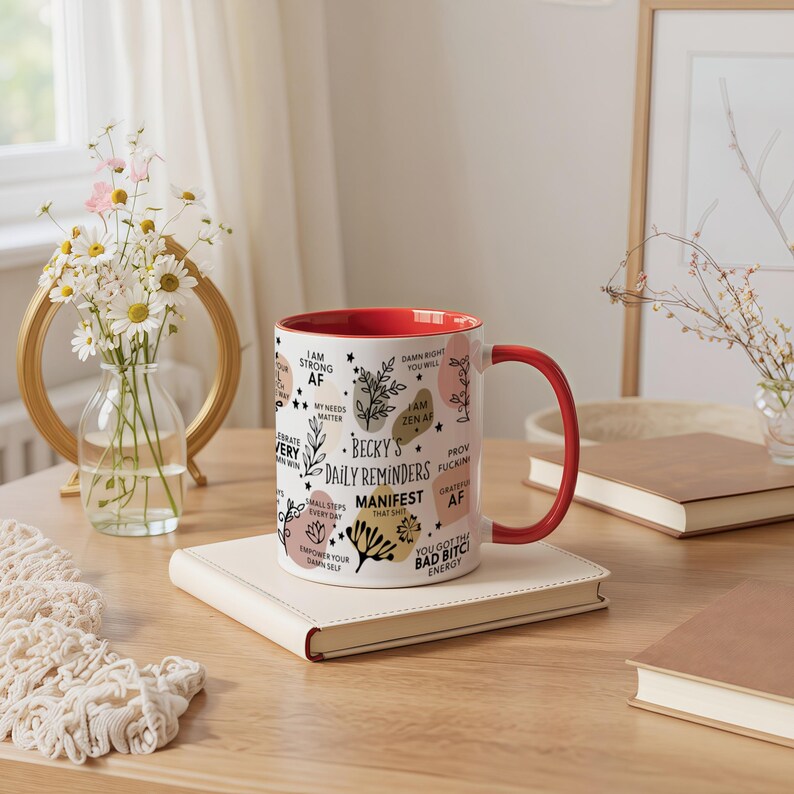 May include: A white ceramic mug with a red interior and handle, featuring the text "Becky's Daily Reminders" and other affirmations. The mug is decorated with floral and abstract designs. It sits on a cream-colored notebook on a wooden table.