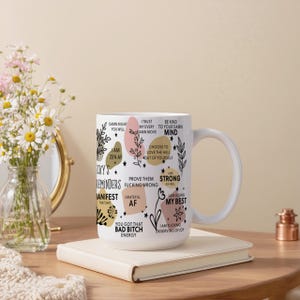 May include: White ceramic mug with a handle, decorated with inspirational phrases and floral accents in black, pink, and gold. The mug features phrases like "Damn Right You Will" and "You Got That Bad Bitch Energy."