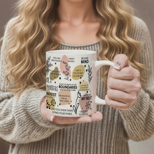 May include: White ceramic mug with inspirational quotes and floral accents. The mug features phrases like "You can and you will," "Be kind," and "I am doing my best." The design includes pastel-colored shapes and botanical illustrations.