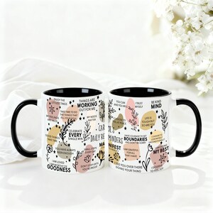 May include: Two white ceramic mugs with black handles and rims. Each mug is decorated with inspirational phrases, floral designs, and colorful shapes. The mugs are on a white surface.