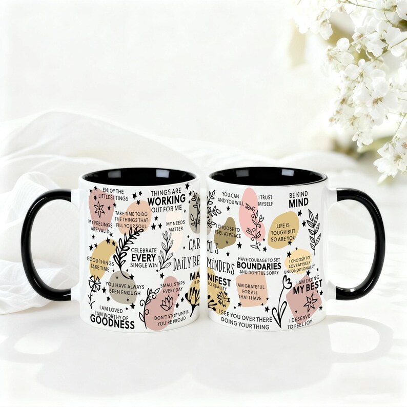 May include: Two white ceramic mugs with black handles and rims. Each mug is decorated with inspirational phrases, floral designs, and colorful shapes. The mugs are on a white surface.