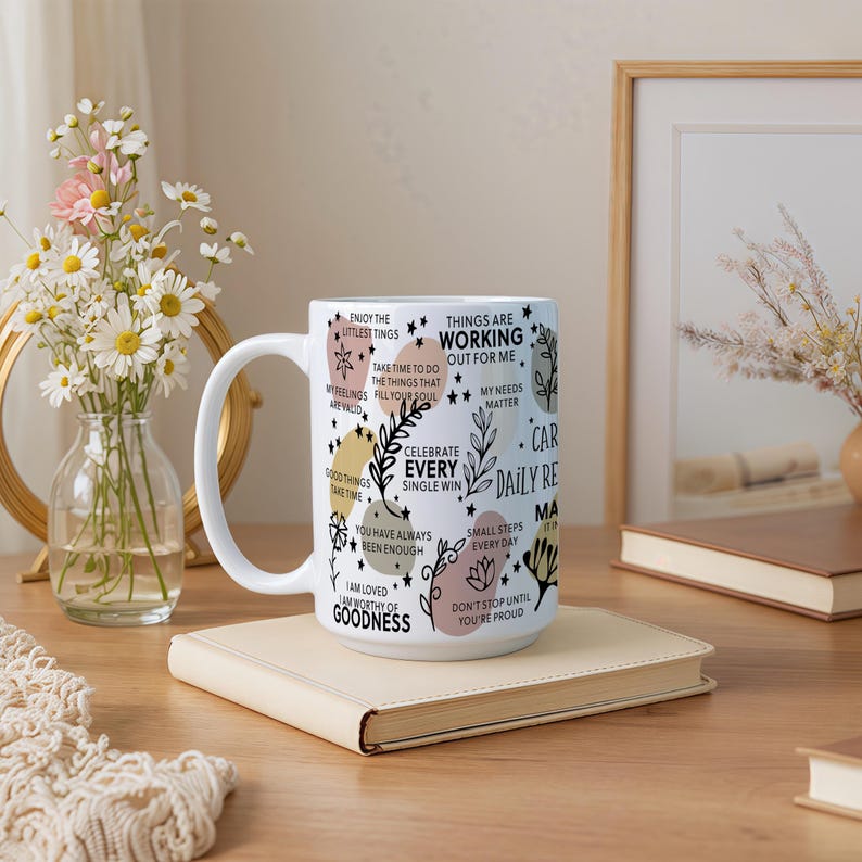 May include: White ceramic mug with a large handle, decorated with inspirational text and floral designs. The mug features phrases like "Celebrate Every Single Win" and "Things Are Working Out For Me". The mug is sitting on a beige book.
