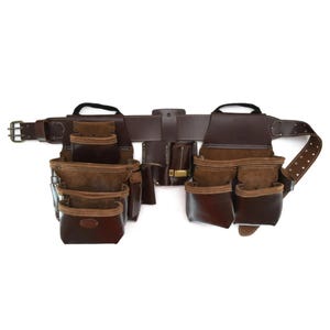 May include: A brown leather tool belt with multiple pockets and pouches. The belt features a buckle closure and is designed for carrying tools. The pockets are reinforced with a lighter brown suede material.