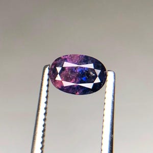 以下が含まれることがあります： 深い紫と青のカラーパレットを持つオーバルカットの宝石で、中央に星のような白い幾何学模様が特徴です。この石は、縁がギザギザになった銀色の金属製ピンセットで保持されています。