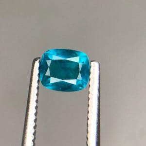 May include: A vibrant, cushion-cut gemstone in shades of teal and turquoise is held by silver tweezers. The gem's facets reflect light, creating a star-like pattern in the center. The background is a soft, neutral gray.
