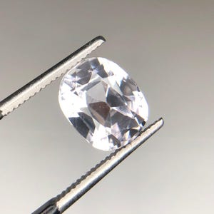 May include: A faceted, oval-shaped, clear gemstone is held by a pair of tweezers. The gem has many reflective surfaces, catching the light and creating a sparkling effect. The tweezers are silver-colored and have a serrated edge.
