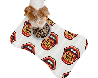 May include: A dog eats from a silver bowl on a bone-shaped pet food mat. The mat is white with a repeating pattern of red lips and the text "EATT THE RICH" in yellow. The dog has brown and white fur.