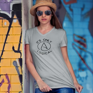 Puede incluir: Una camiseta gris claro con cuello en V con el texto "IT'S ONLY LOGICAL" en negro. La camiseta presenta un diseño circular con la letra "A" en su interior. La persona lleva vaqueros azules y un sombrero de paja con gafas de sol.