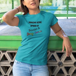 May include: Teal t-shirt with the text "IMAGINE BEING AFRAID OF BOOKS & Drag Queens" in black and rainbow colors. The shirt is paired with light blue jeans. The person is leaning against a green and gray wall.