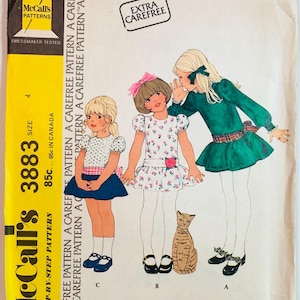 May include: Vintage McCall's 3883 sewing pattern for a child's dress. The pattern features illustrations of three girls in dresses, with a cat illustration. The pattern is marked "Extra Carefree" and "Dressmaker Tested".