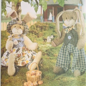 May include: A vintage Butterick pattern cover featuring two plush bunny dolls, Hedda and Henry Hare, in a garden setting. Hedda wears a floral dress and hat, while Henry sports plaid pants and a vest. A basket of apples sits nearby.