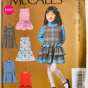 May include: McCall's sewing pattern M7008 for girls' dresses. The pattern features various dress styles, including a pink gingham dress with a bow, a floral dress, and a denim dress. The cover shows a girl wearing a plaid dress with blue bows.