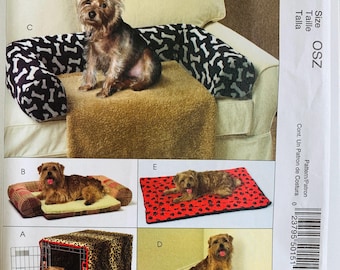 May include: A sewing pattern envelope for pet beds and crate covers featuring five variations labeled A through E. The images display dogs on a leopard print crate cover, a plaid bolster bed, a bone-patterned sofa guard, a corner mat, and a paw print mat.