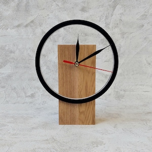 May include: A modern desk clock with a black circular frame and a wooden base. The clock features black hour and minute hands, and a red second hand. The wooden base is rectangular and has a natural wood grain finish.