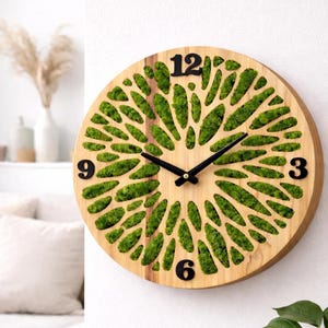 Op de afbeelding: Een ronde houten wandklok met een natuurlijke houtnerf. De wijzerplaat heeft een patroon van groen mos en zwarte cijfers op de posities 3, 6, 9 en 12 uur. De wijzers zijn zwart.