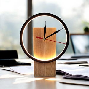 Puede incluir: Un reloj de escritorio moderno con un marco negro circular y una base de madera. La esfera del reloj es un bloque de madera con manecillas negras y rojas. El reloj está iluminado desde dentro, creando un brillo suave.