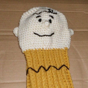 May include: A crocheted golf club cover featuring a smiling cartoon character with black eyes and a black zig-zag line across the bottom. The character's head is white and the body is yellow.