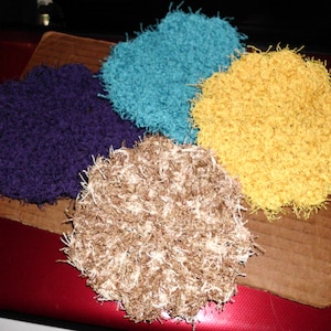 May include: Four crocheted coasters in different colors: purple, teal, yellow, and brown. The coasters are round and have a fluffy texture.