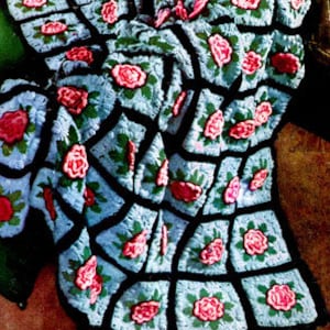 May include: A crocheted blanket with a repeating pattern of pink roses and green leaves on a blue background. The blanket is made up of squares with black borders.