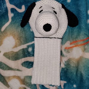 Könnte beinhalten: Eine gehäkelte Schlägerhaube in Weiß und Schwarz, die einer Zeichentrickfigur ähnelt, mit schwarzen Ohren und einer schwarzen Nase. Die Hülle hat einen langen, rechteckigen weißen Körper.