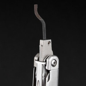 May include: A multi-tool component featuring a silver-toned metal body with a textured finish and a single, curved, dark grey metal hook extending upwards from the centre. The tool includes a visible screw head on the side and a hinge mechanism.