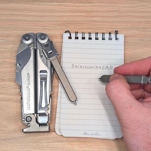 May include: A silver Leatherman Surge multi-tool and a small notepad with the text "Backcountry CAD" written on it. A hand is holding a pen and writing on the notepad. The notepad is labeled "Rite in the Rain".