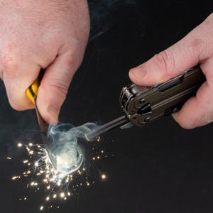 May include: A multi-tool with a dark metallic finish and a ferrocerium rod is being struck by a yellow-handled tool, creating a bright shower of sparks and a thin trail of white smoke against a solid black background.
