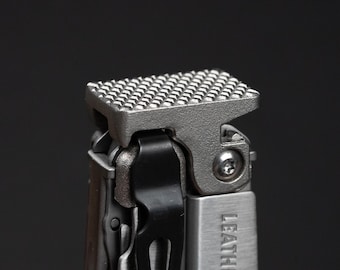 May include: A silver-toned multi-tool featuring a textured, cross-hatched hammer surface at the top. A black metal pocket clip is attached to the side, and the word LEATHERMAN is partially visible in embossed lettering on the brushed metal handle.