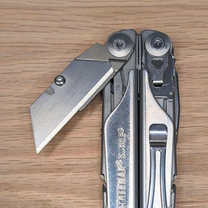 May include: A multi-tool with a partially extended blade. The tool is silver and has the text "LEATHERMAN SURGE" on the side. The blade is angled and sharp, with a silver finish. The background is a light brown wooden surface.