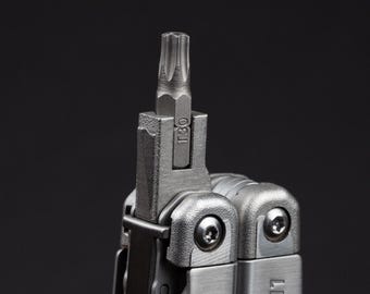 May include: A silver-colored multi-tool featuring a T30 Torx bit extended from the main body. The metal tool has a brushed finish with visible screws and the partial brand name LEATH visible on the side of the handle against a dark background.