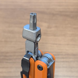 May include: A multi-tool with an orange handle and a silver metal tool bit extended. The tool has a black pocket clip and several other tools folded inside. The bit is a star-shaped Torx head.