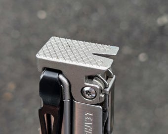 May include: Close-up of a silver multi-tool with a textured top and a black clip. The tool has the words "LEATHERMAN SURGE" printed on the side. The top of the tool has a notch and a textured surface.