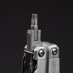Peut inclure: Un outil multifonction argenté doté d'un embout Torx T30 inséré dans la fente centrale. L'outil est fabriqué en métal brossé avec des vis apparentes et le nom de marque partiel LEATH visible sur le côté du cadre de la poignée.