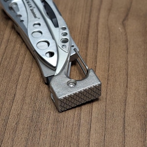 May include: A close-up of a silver multi-tool with a textured hammer head and a carabiner clip. The tool is partially open, revealing other components. The tool is on a wooden surface.