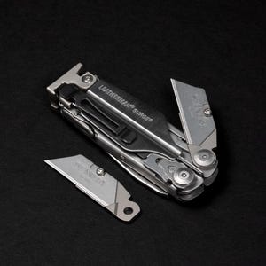 Può includere: Un utensile multifunzione Leatherman Surge color argento con una clip da tasca nera e una lama Stanley fissata. Una lama Stanley color argento abbinata è posizionata accanto all'utensile. Entrambe le lame riportano il marchio Stanley.