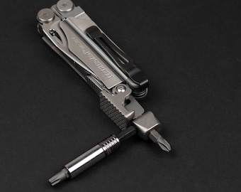 Può includere: Un utensile multifunzione Leatherman Wave+ color argento in posizione ripiegata con una clip da tasca nera. Un portainserti in metallo è esteso e sostiene un inserto per cacciavite a doppia estremità con testa a croce e Torx.