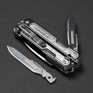 May include: A multi-tool with a topographic pattern on the handle, with a partially opened blade and an additional blade lying next to it. The multi-tool is silver and black, with the brand name "LEATHERMAN ARC" visible on the handle.