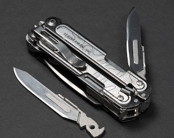 May include: A multi-tool with a topographic pattern on the handle, with a partially opened blade and an additional blade lying next to it. The multi-tool is silver and black, with the brand name "LEATHERMAN ARC" visible on the handle.